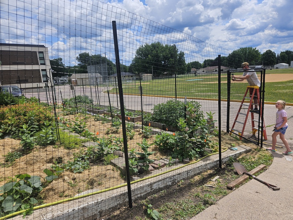 The Middle School garden with the new fence being worked on, 1 person on a ladder and the other person carrying pieces.
