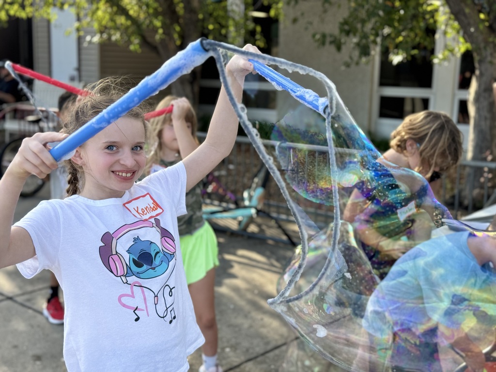 A girl wearing a white shirt holds a giant bubble wand in front of her. Other children play in the background.