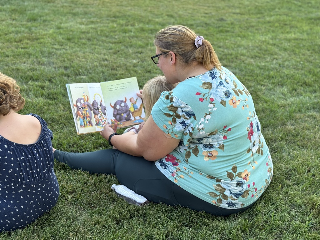 A woman holds a book and reads aloud to a child on the lawn.