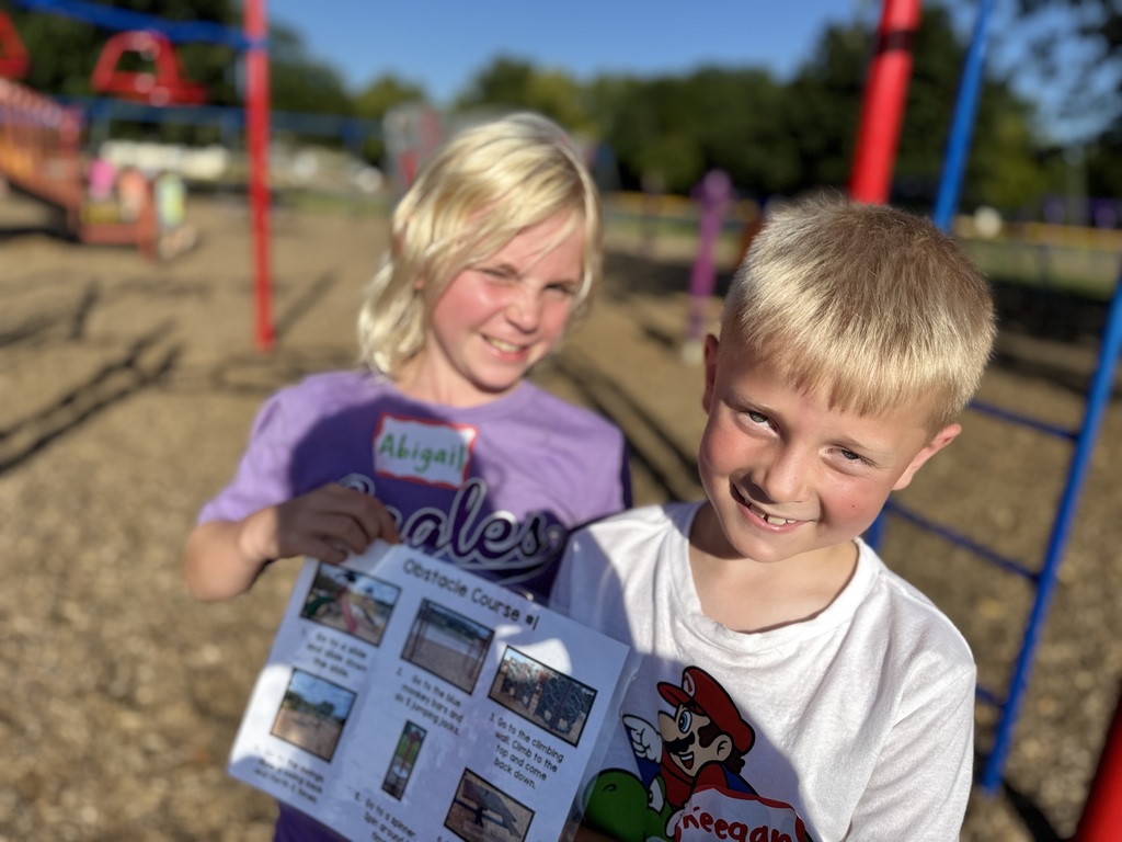 Two kids stand by the playground, one holds a flyer with the words "obstacle course" while the other smiles.