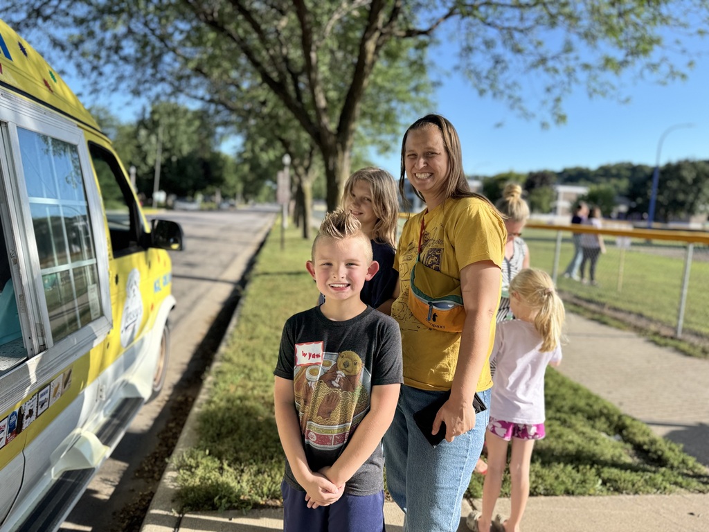 Family with two kids stands by a yellow ice cream van on a sidewalk, with a grassy area and trees in the background.