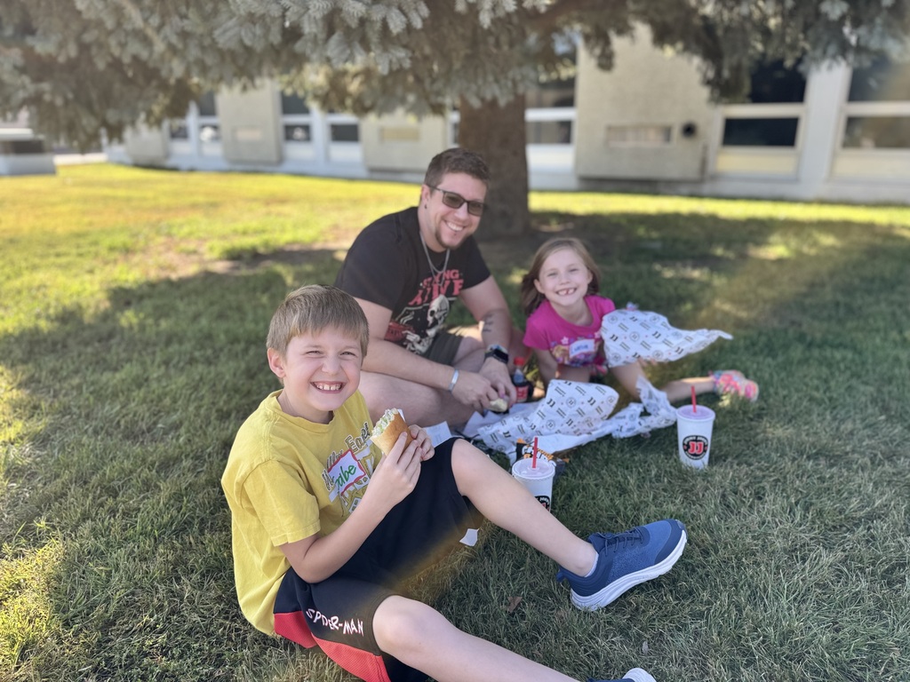 Family sitting in a grassy area, one boy eating, one girl sitting, and a man in sunglasses.