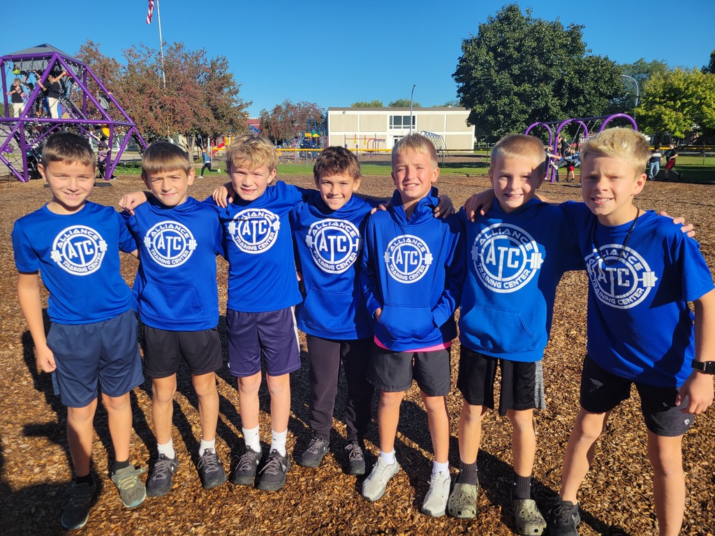 7 young boys in blue shirts with white letters pose together at a playground, with trees in the background.