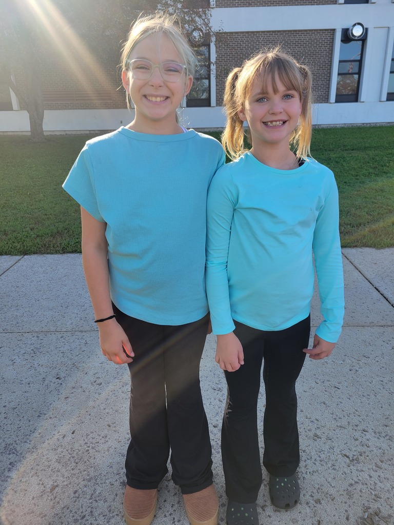Two young girls in teal shirts and black pants standing outside, smiling, on a concrete sidewalk.