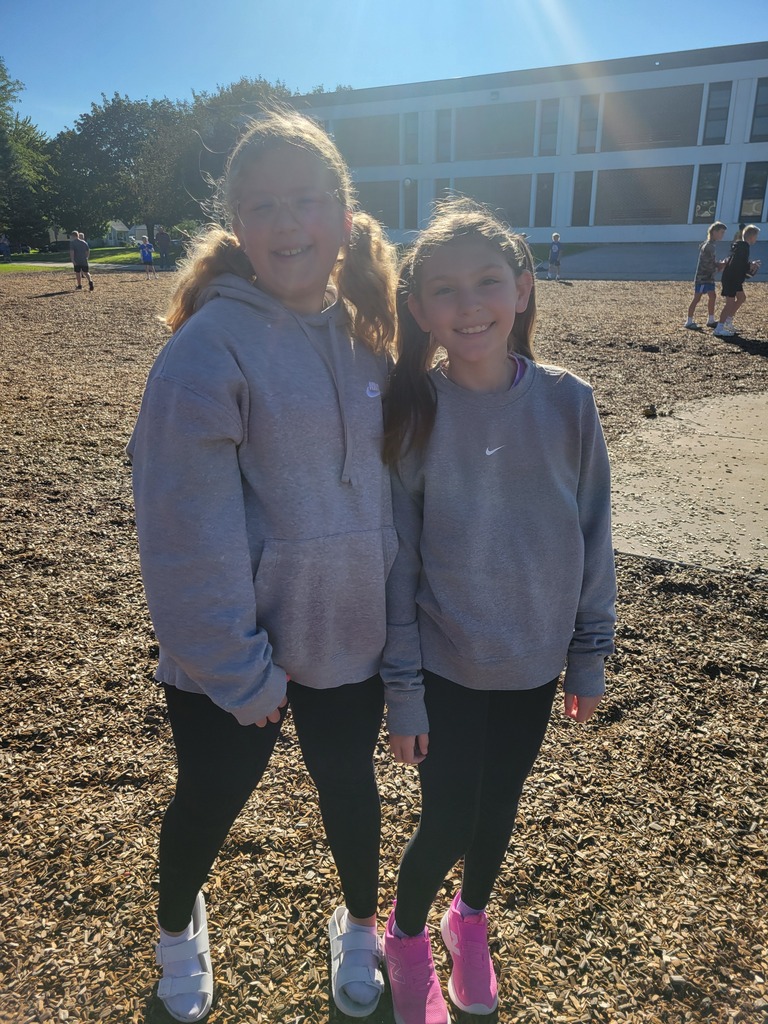 Two girls pose close together on a field in front of a building, both wearing gray sweatshirts and black leggings.