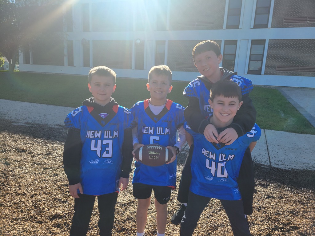 Four boys in blue jerseys pose for a photo outside. They hold a football between them.