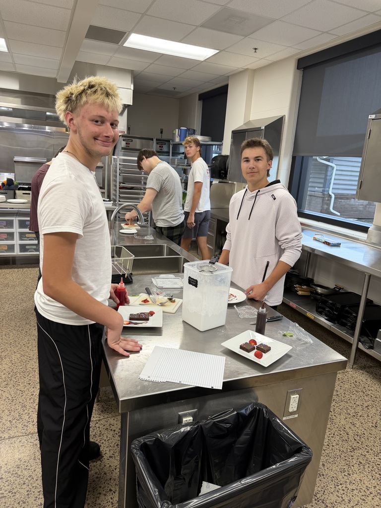 Two students in white shirts stand in a kitchen working on their plates.