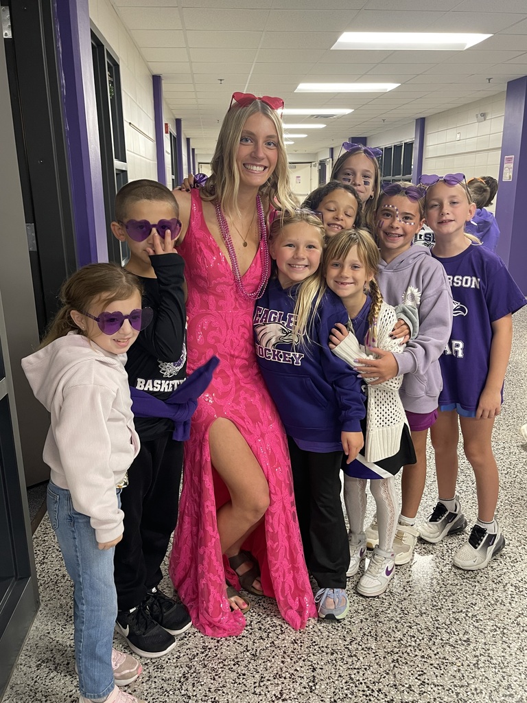 A homecoming court student in a pink dress stands with children in purple and white, wearing sunglasses, in a school corridor.