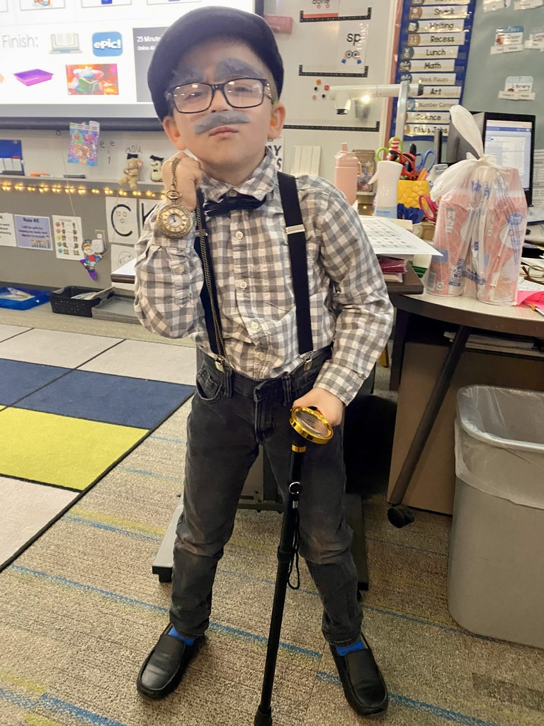An elementary student is dressed up for the 100th day of school.  He's holding his cane, and magnifying glass in one hand and a watch on a chain in the other hand. He is wearing a fake grey mustache and eyebrows. 