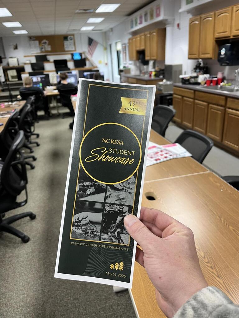 A hand holds a black and gold program for the “NC RESA Student Showcase,” labeled “43rd Annual,” in the foreground. The background shows a classroom or workspace with several people seated at computers, a long counter with wooden cabinets, and a kitchenette area. The program also includes the location “Dogwood Center of Performing Arts” and the date May 14, 2026.