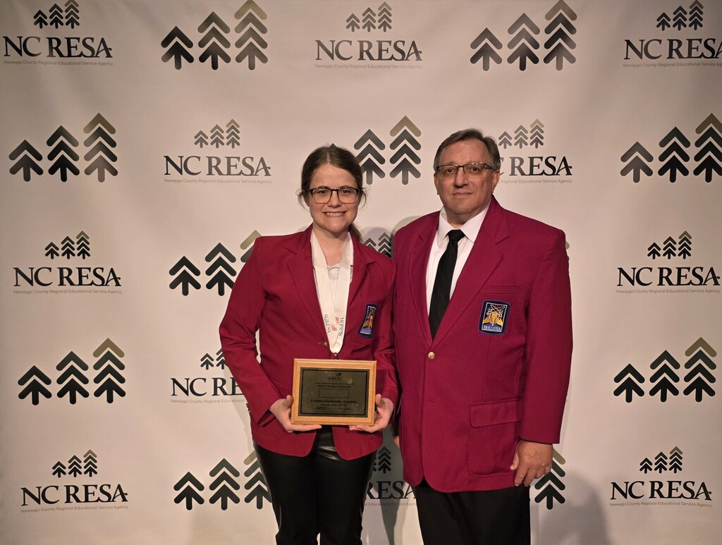 Two individuals in red FFA jackets stand together holding an award plaque in front of a display backdrop.