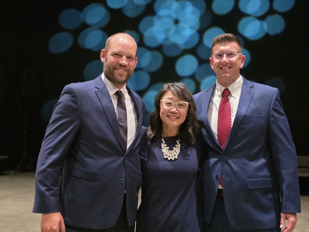 Three individuals in formal attire pose together for a photo on stage at the showcase.