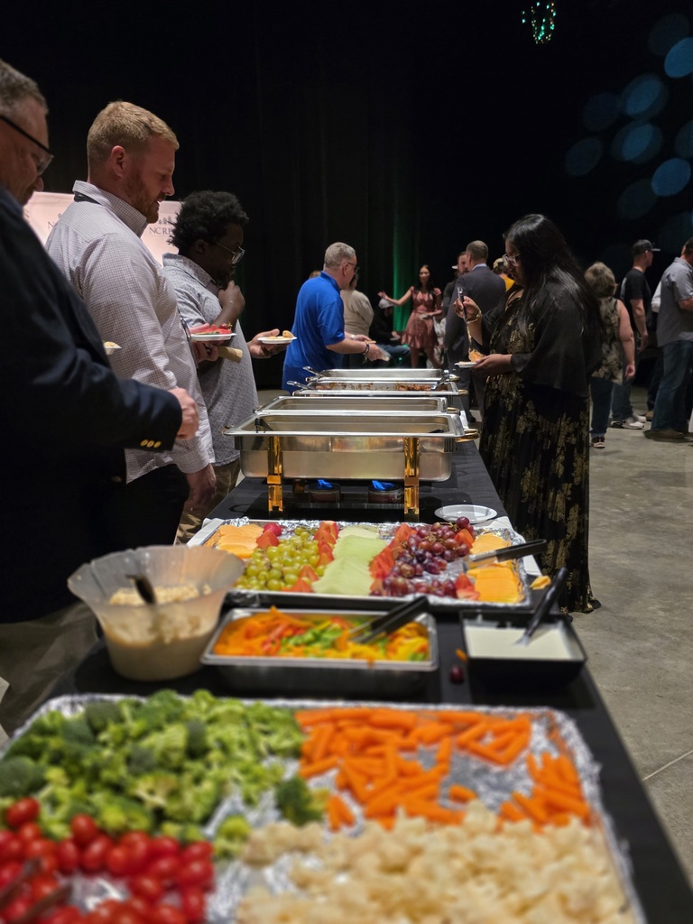 Guests stand in line serving themselves food from a buffet table with fruits, vegetables, and hot dishes at the NC RESA Student Showcase.
