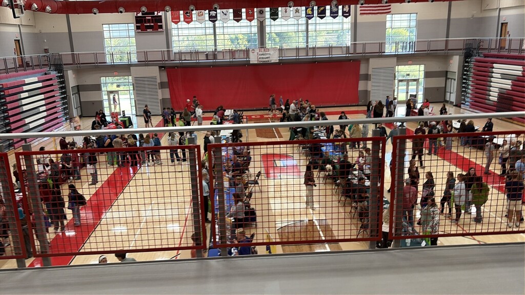 An indoor basketball court with a crowd of people seated and standing, red-floored, and a red wall.