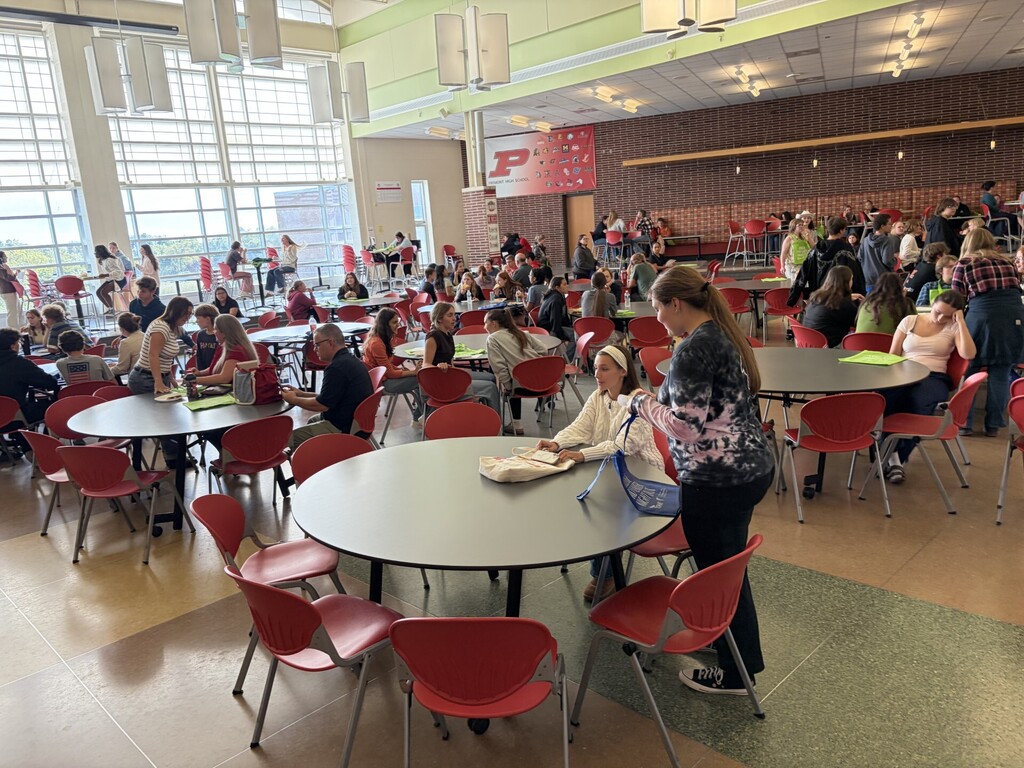 Large cafeteria with people seated around tables. Large windows allow natural light to fill the space.