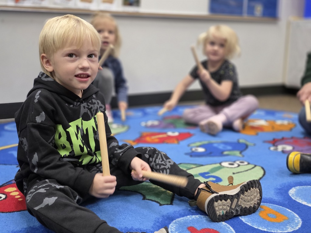 Children in a classroom with a blue carpet, one holding drumsticks and another playing a drum.