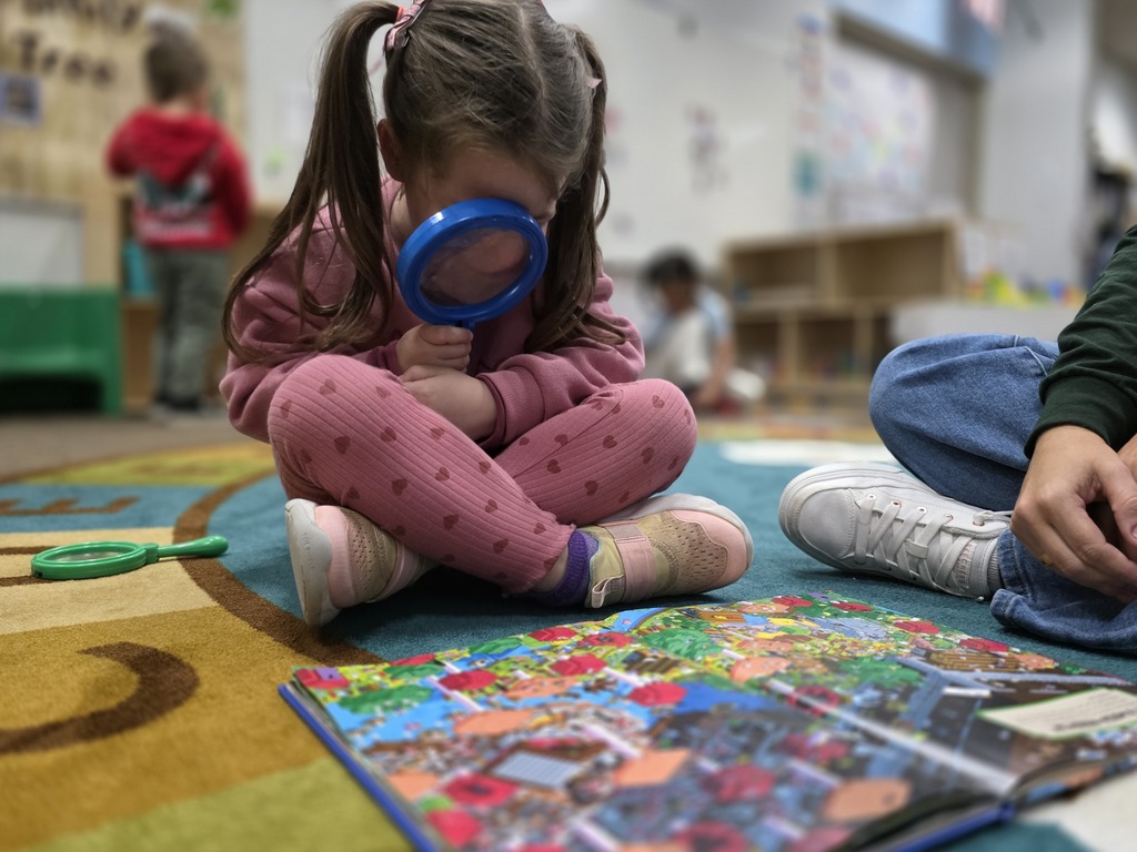 A child with a magnifying glass examines a colorful book on a colorful floor while seated.