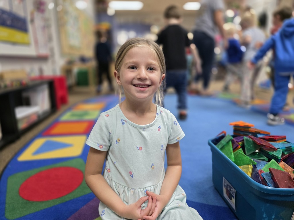 A smiling girl in a white dress sits on a colorful carpet in a classroom. Behind her, other children walk around.