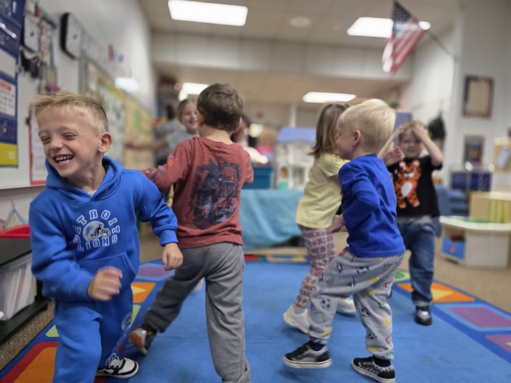 Young children wearing various colored hoodies and pants dance and laugh in a brightly lit classroom.