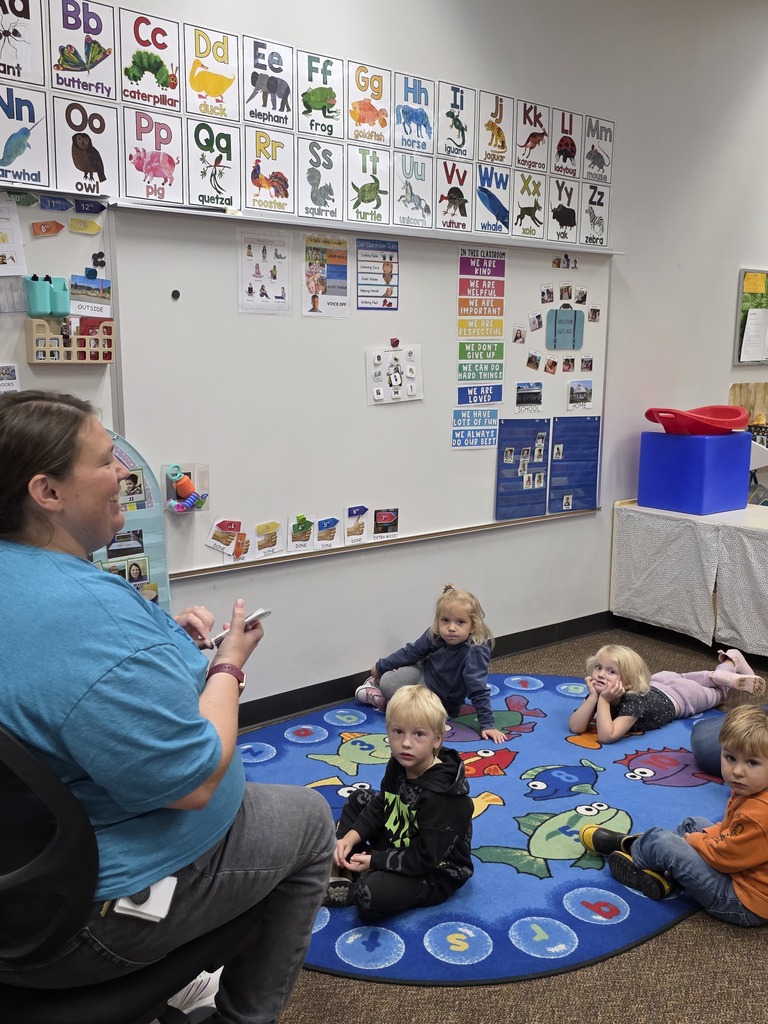 Woman sits with children on rug in classroom, speaking to them. Alphabet charts, toys, and desk with tablecloth.