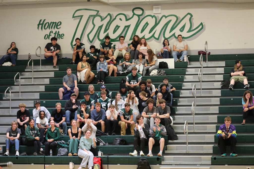 Students on the bleachers with the "Home of the Trojans" logo above them on the wall