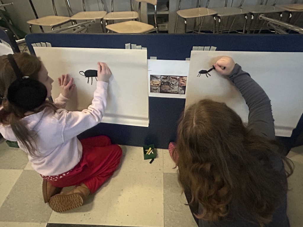 Two students work on their cave inspired paintings, sit on floor in front of easel.