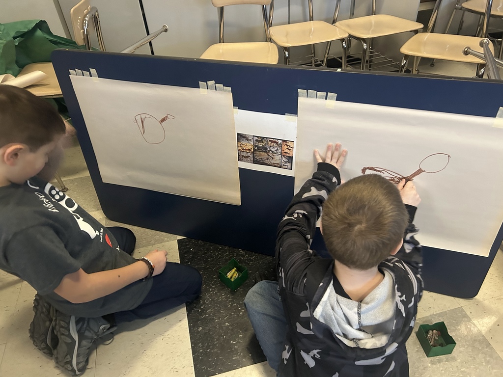 Two students work on their cave inspired paintings, sit on floor in front of easel.
