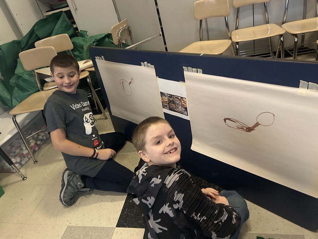 Two students work on their cave inspired paintings, sit on floor in front of easel.