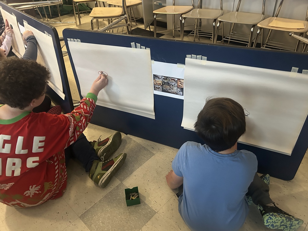 Two students work on their cave inspired paintings, sit on floor in front of easel.