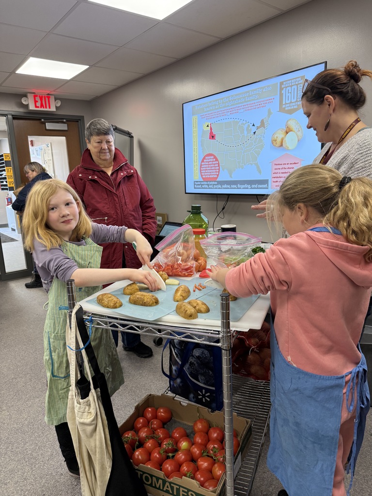 Elementary students participate in a Kids Farmers Market cooking demo, cutting potatoes on a table while wearing aprons. Fresh produce including potatoes, tomatoes, onions, and herbs is displayed nearby. Adults stand behind the students observing and offering guidance during the hands-on activity.