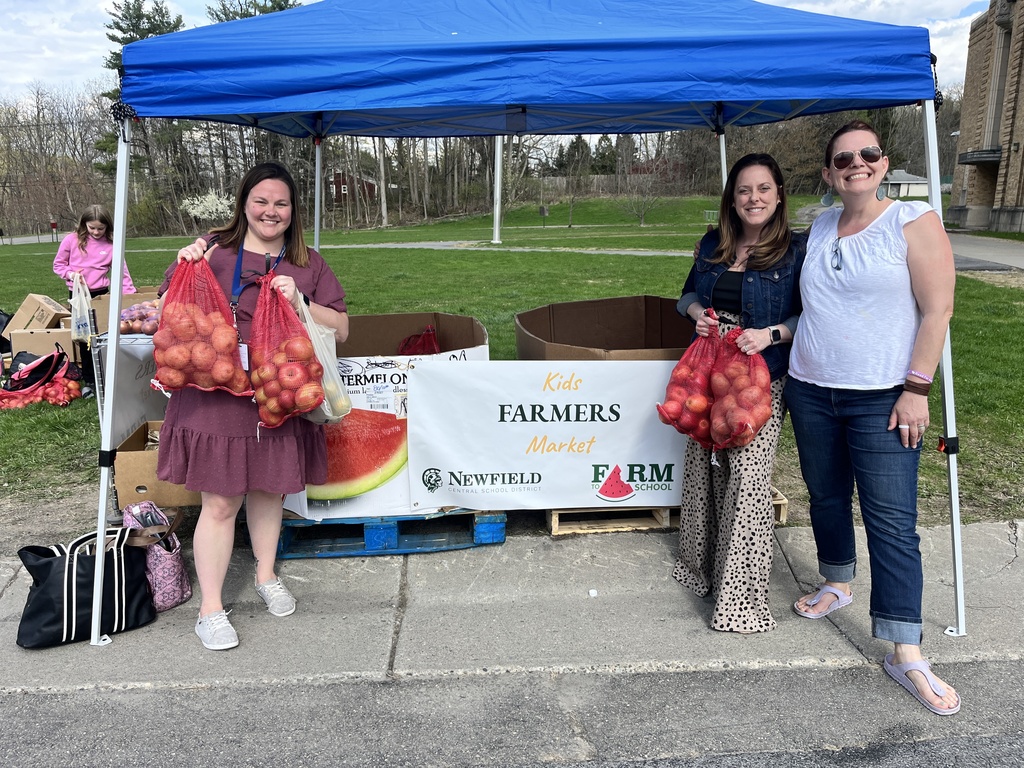 Three adults stand under a blue pop-up tent at a Kids Farmers Market, smiling and holding bags of apples. Boxes of produce are stacked behind them, and a Kids Farmers Market banner with Newfield Central School District and Farm to School logos is displayed at the front. The market is set up outdoors on a grassy school campus.