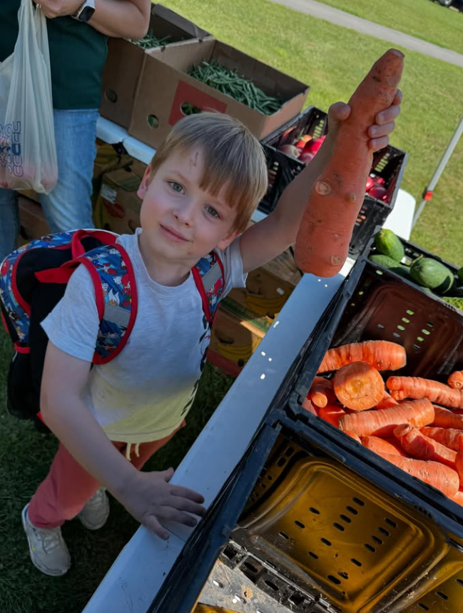 Child wearing a backpack smiles and holds up a large carrot at an outdoor Kids Farmers Market. Crates filled with carrots and other fresh vegetables are displayed on tables nearby.