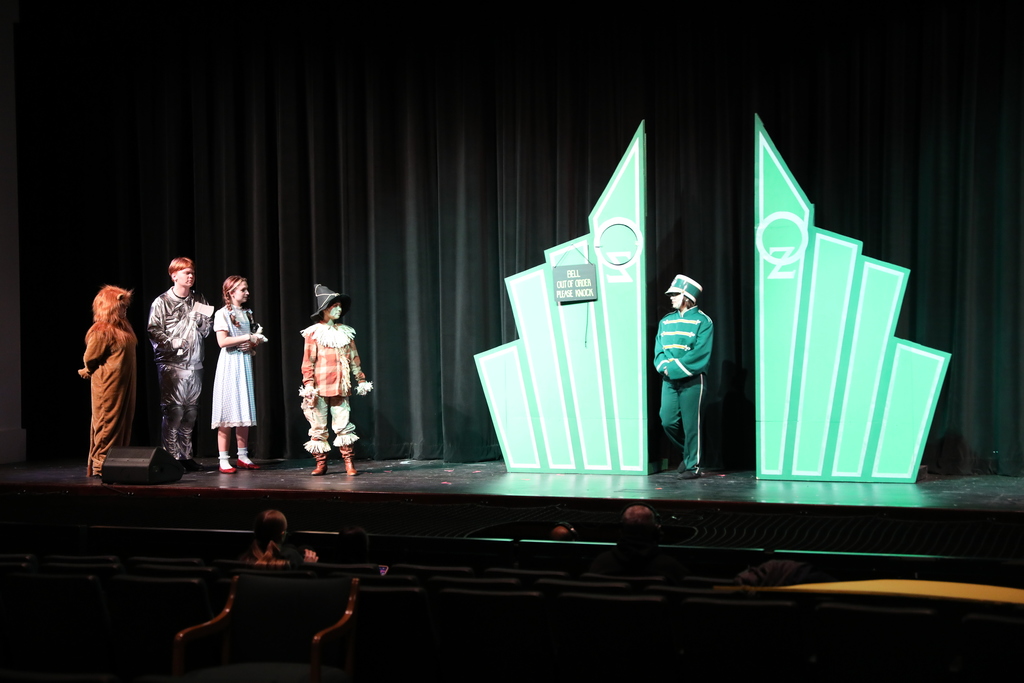 Students perform a scene from The Wizard of Oz on a school stage. Characters including the Cowardly Lion, Tin Man, Dorothy, and Scarecrow stand facing a guard at the glowing green gates of the Emerald City, with a dark theater audience visible in the foreground.
