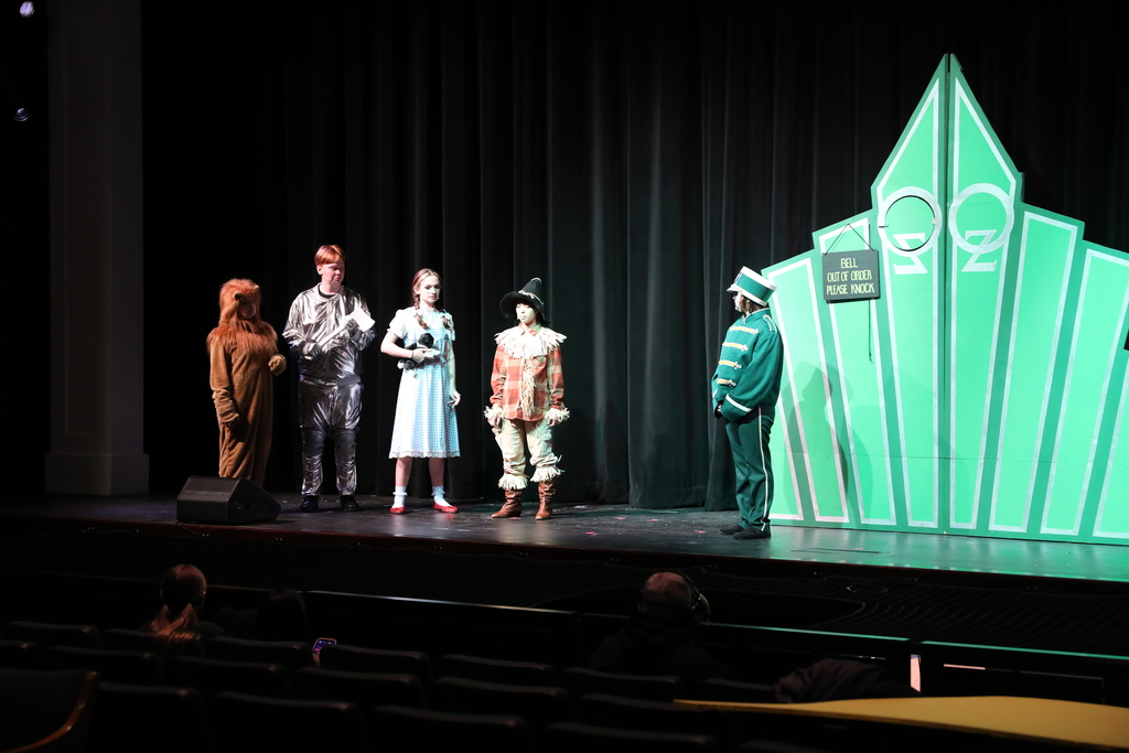 Students perform a scene from The Wizard of Oz on a school stage. Characters including the Cowardly Lion, Tin Man, Dorothy, and Scarecrow stand facing a guard at the glowing green gates of the Emerald City, with a dark theater audience visible in the foreground.