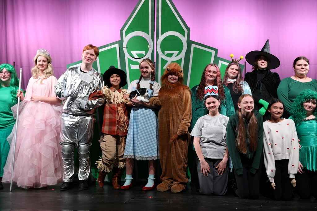 Group of students in costume pose on stage in front of a green Emerald City backdrop during a production of The Wizard of Oz. Characters include Glinda, the Tin Man, Scarecrow, Dorothy holding Toto, the Cowardly Lion, and the Wicked Witch, along with other cast members dressed in green-themed outfits.