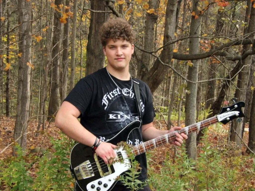 A student standing in front of a forest will holding a bass guitar and posing for a picture for a Tompkins Weekly article about him