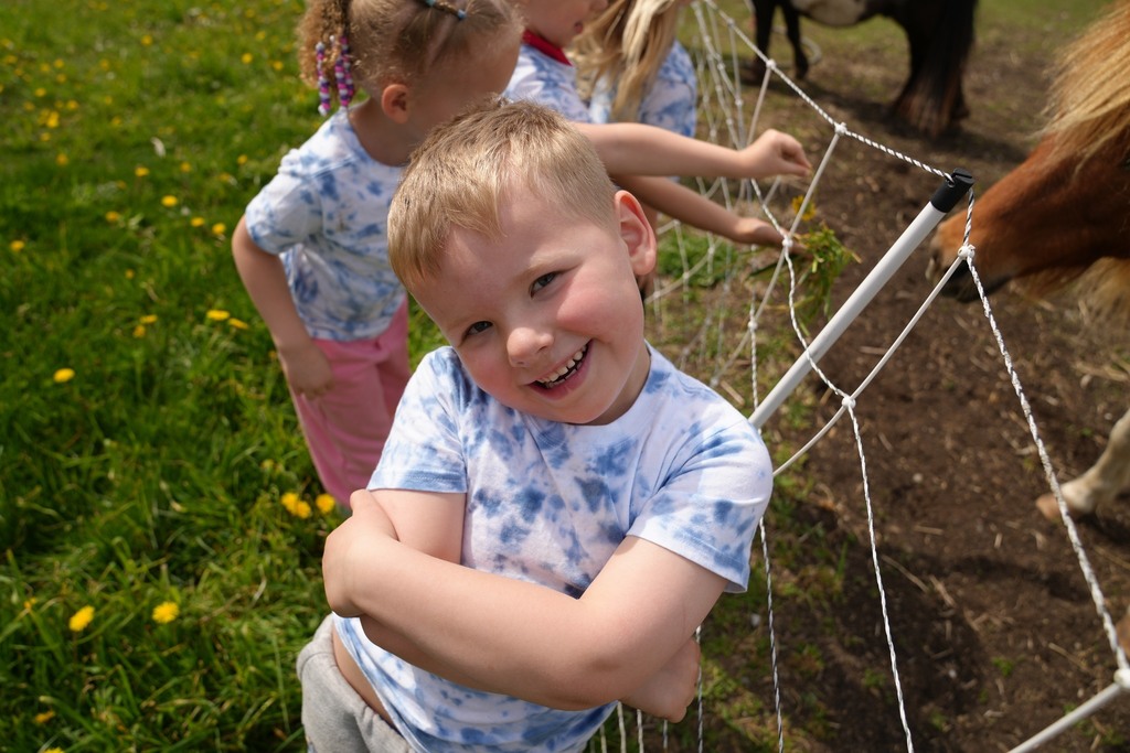 Pre-K student smiling with arms crossed while standing near a fence feeding miniature horses at Daisy Hollow Farm.