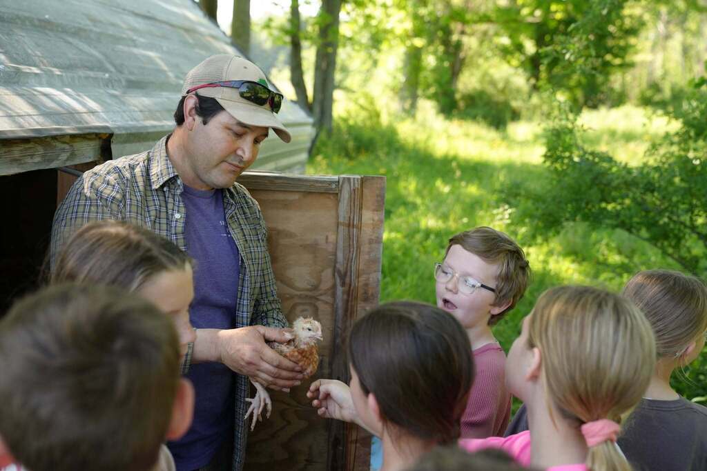 A farmer shows students a young chicken while explaining poultry care and farm operations during a field trip.