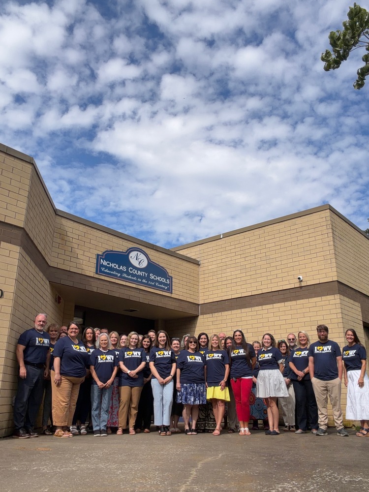 Group photo of approximately 30 Nicholas County Schools administrators and staff standing outside the entrance of a tan brick school district building. A sign above the doorway reads, “Nicholas County Schools – Educating Students in the 21st Century.” The group is gathered on the sidewalk in front of the building, many wearing matching navy blue “I ♥ WV Public Schools” T-shirts. The photo is taken from a low angle, showing a large blue sky filled with scattered white clouds above the building. The image represents district leadership and staff coming together at the start of the new school year.