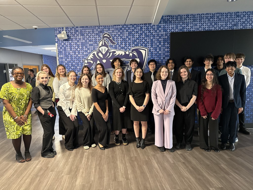 Group of people in formal attire standing in a room with blue walls and wooden flooring, smiling.
