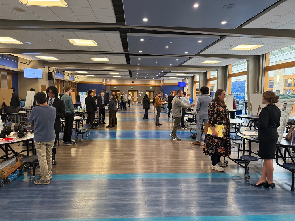 Indoor event with tables, people in a hallway, some standing and some walking, blue floor with white lines.