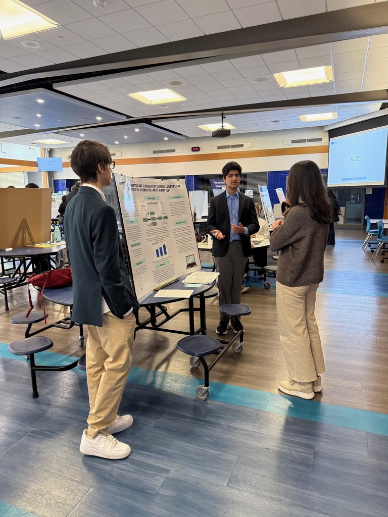 Three people in suits stand by a table with a presentation board. A woman in white shoes listens while a man speaks.