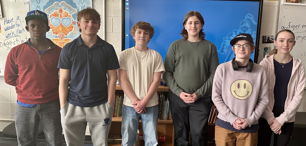 Five young individuals stand in a classroom, smiling, with a blue screen in the background. Behind them are shelves with books and artwork.