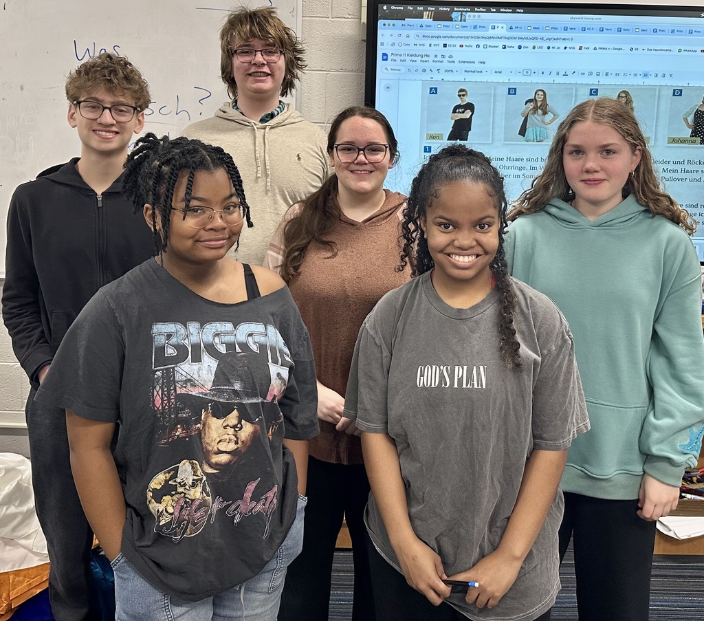 Six young people standing together. Three girls wearing glasses. A monitor showing pictures of people.