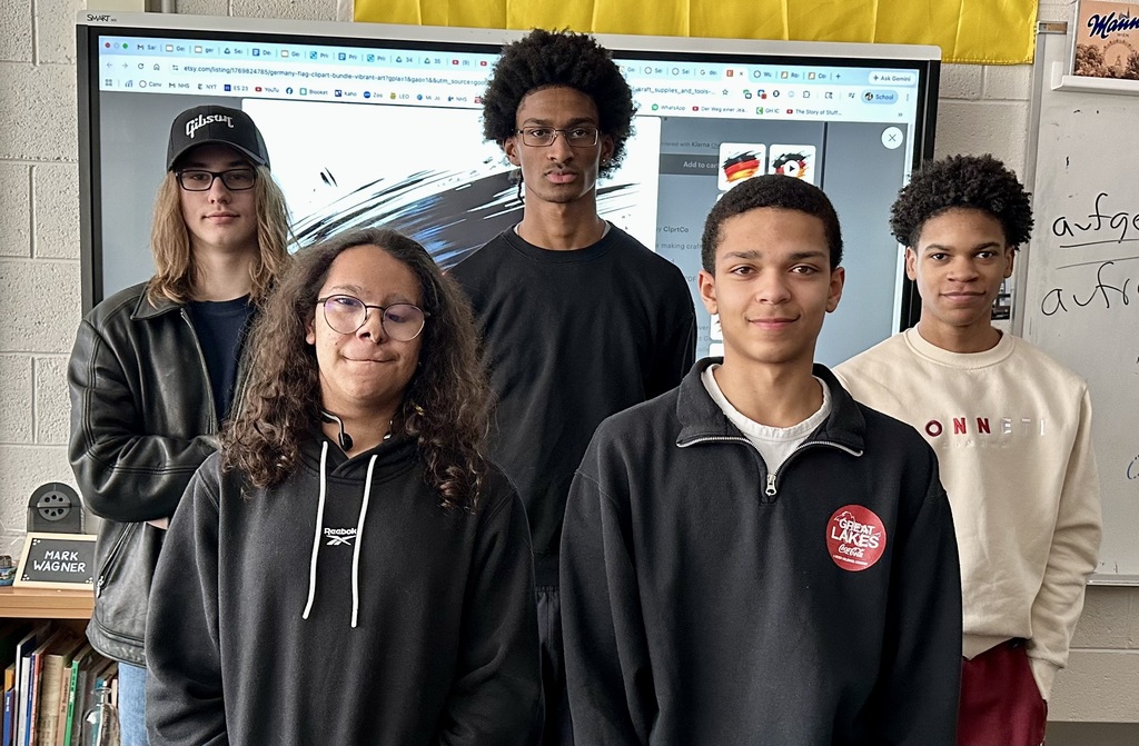 Five individuals standing together in a classroom, smiling. Behind them, a large monitor and a whiteboard.