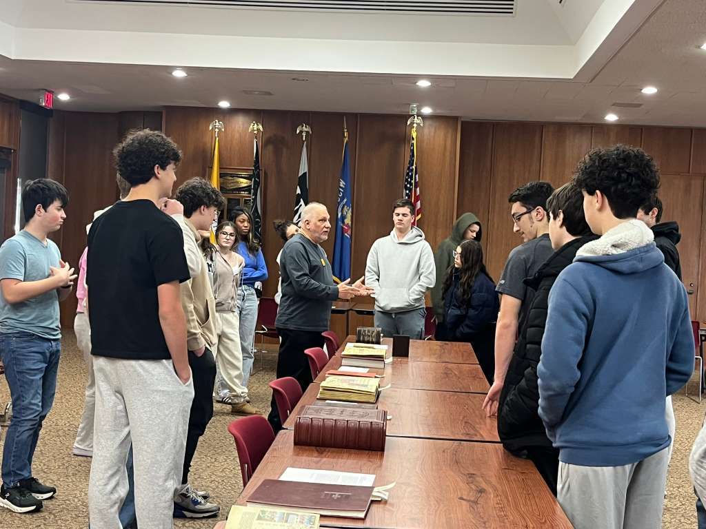 Group of people in a room with a long table, books, and flags behind the man in the center.