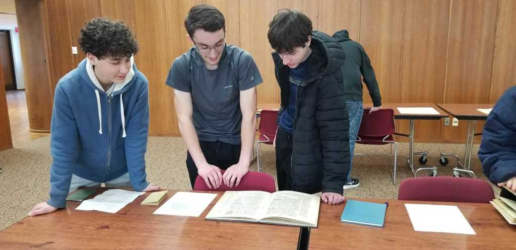 Three people in a classroom, focused on a table with books and papers. Behind them, a person stands near a desk.