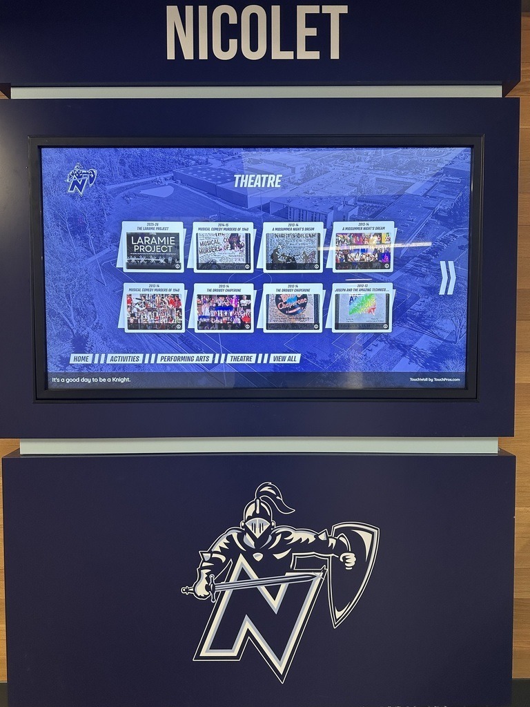 A blue kiosk displays a screen with "NICOLET" and "THEATRE" on top. The screen shows various images. Below, a knight emblem reads "N".