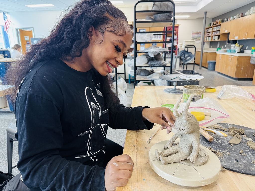 Person sitting at a table, creating a clay figure. Background contains tables, shelves, and materials.