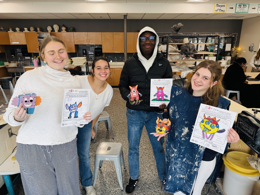 Four individuals in a room with cabinets and stools, holding up crafted paper toys and smiling.